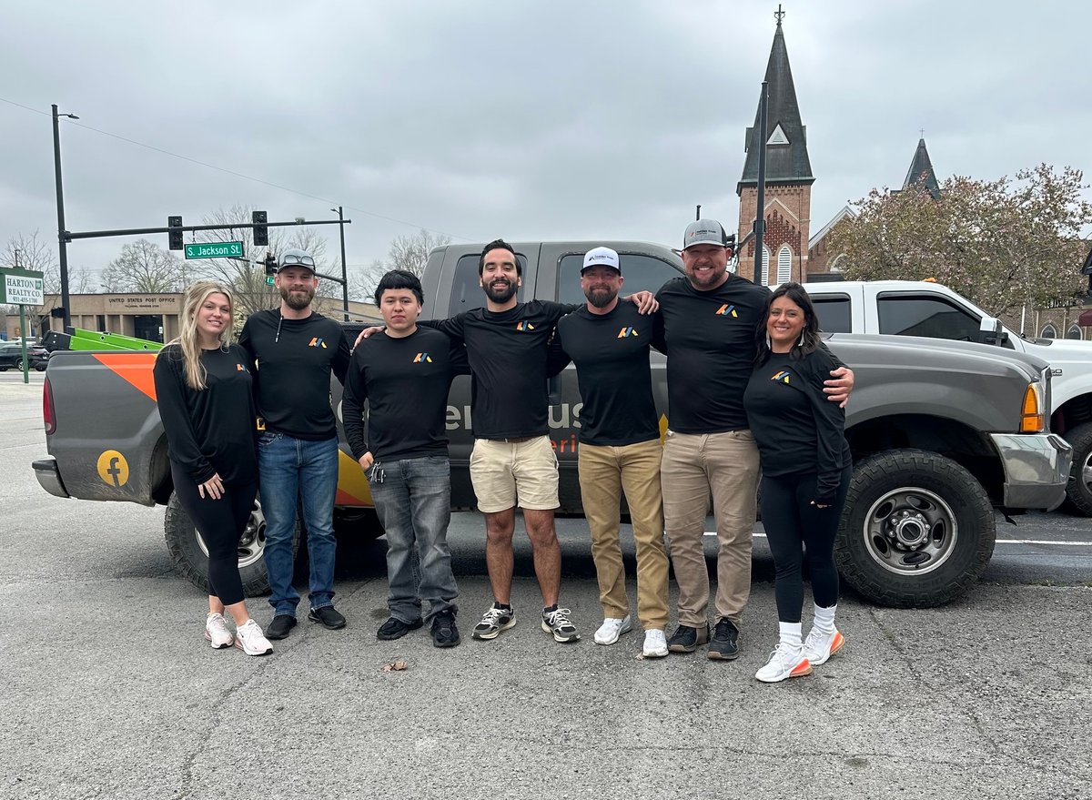 Group of people in black shirts posing in front of a black truck with a rainbow graphic, with a church tower visible in the background