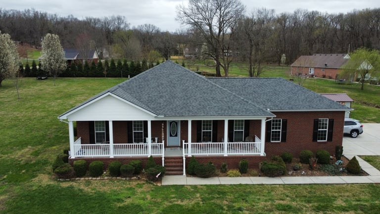 Red brick ranch house with white front porch and columns, gray shingle roof, on green lawn with bare trees in background