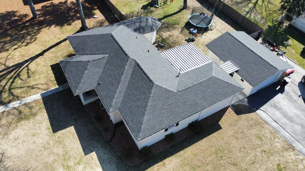 Aerial view of a residential house with gray shingle roof, white gutters, and driveway surrounded by trees and grass yard
